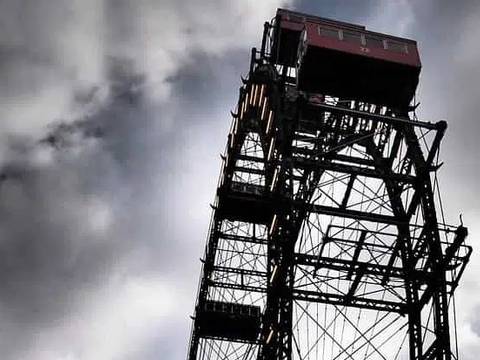  – Großes Riesenrad in metallischer Struktur vor wolkigem Himmel. Oben rechts eine rote Gondel, unten links ein beleuchtetes Kreuz auf einem Dach.
