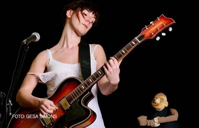 PAUL UND PAULA - EINE LEGENDE – Person spielt E-Gitarre neben Mikrophon. Die Person trägt ein Trägerkleid. Daneben eine Puppe in schwarzem Hemd. Text: FOTO: GESA SIMONS.