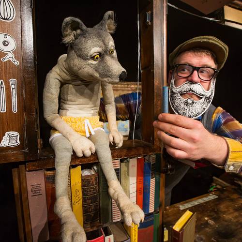 Wolfspuppe in gelben Shorts sitzt auf einem Regal mit Buechern. Person mit Brille und Papierbart haelt das Regal.