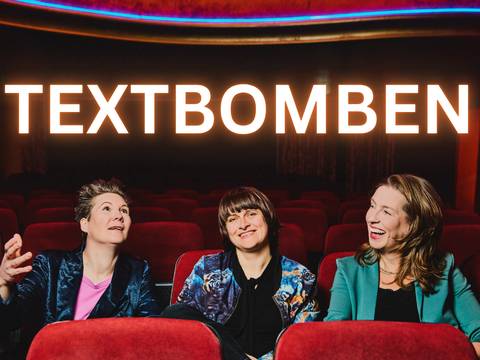 Die Textbomben – Drei lachende Personen in einem Theater mit roten Sitzen, darüber leuchtender Schriftzug "TEXTBOMBEN".