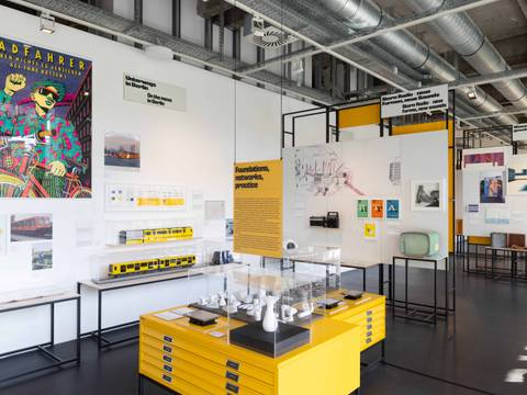 Ausstellungsansicht „Gestalten für Berlin. Design aus der Kunsthochschule Berlin-Weißensee“ – Ausstellungsbereich zum Thema Mobilität in Berlin mit Modellen von U-Bahnen in Vitrinen, einem bunten Plakat 'Radfahrer' und verschiedenen Designobjekten auf gelben Podesten