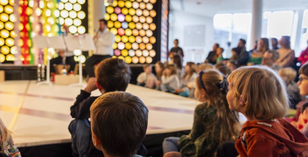 Eine Gruppe von Kindern sitzt auf dem Boden vor einer Bühne, auf der zwei Personen musizieren. Im Hintergrund leuchtet eine bunte Wabenwand.
