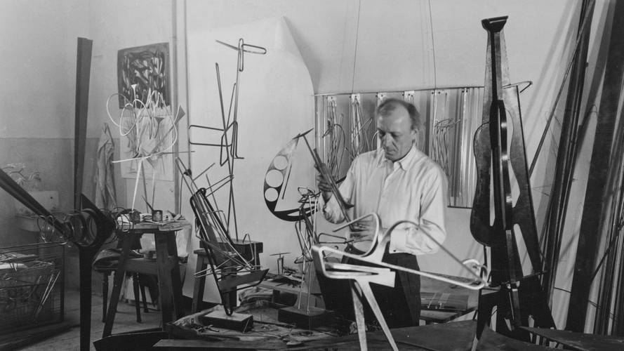 Ewald Gnilka, Ohne Titel (Hans Uhlmann in seinem Atelier), um 1954 – Schwarz-Weiss-Fotografie von Hans Uhlmann in seinem Atelier. Um den Künstler herum sind viele Metall-Skulpturen im Entstehungsprozess zu sehen.