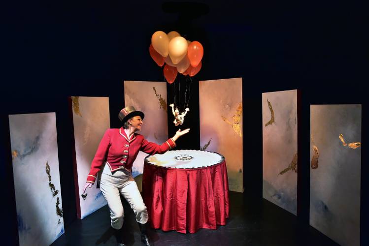 Person in roter Jacke und Hut hält Luftballons. Tisch mit rotem Tuch, umgeben von bemalten Leinwänden.