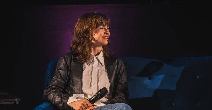 Lucilla Schmidinger – Person sitzt auf blauem Sofa, hält Mikrofon in der Hand, trägt schwarzes Lederjackett und weißes Hemd, lächelt, violettes Licht im Hintergrund.