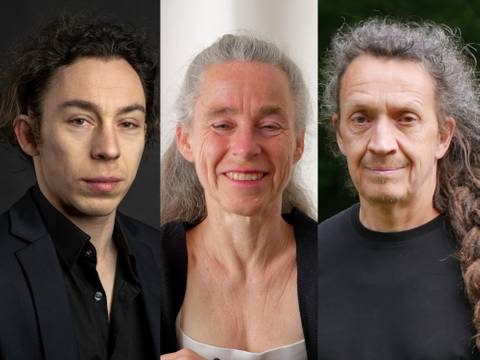  – Drei Personen nebeneinander: links mit Anzug, Mitte lächelnd mit grauen Haaren, rechts mit langen Locken und schwarzem T-Shirt.