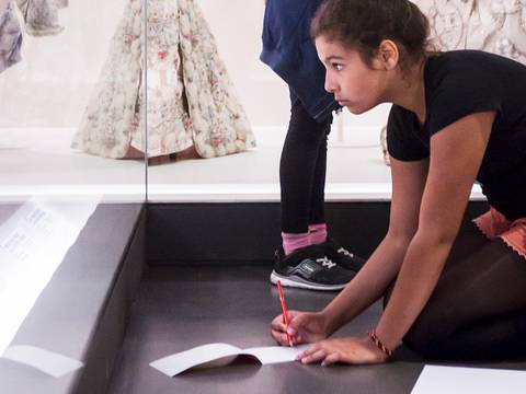  – Kind kniet auf Boden mit rotem Stift und Papier vor einer Vitrine mit historischen Kleidern im Museum. Wahrnehmung konzentriert auf Notizen.