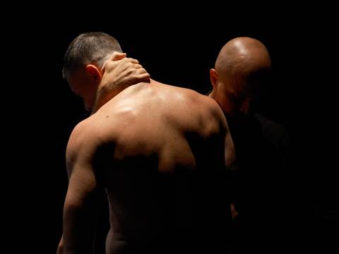 THE SHAPE OF A MAN – Zwei Personen im Dunkeln; eine mit freiem Oberkörper, schaut nach unten. Eine Hand berührt den Nacken der anderen Person, die sich teilweise im Schatten