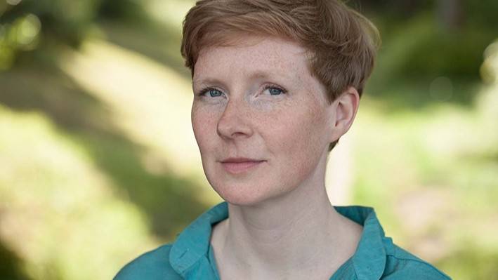 Judith Zander – Person mit kurzen Haaren im türkisfarbenen Hemd blickt in die Kamera; unscharfer, grüner Hintergrund.