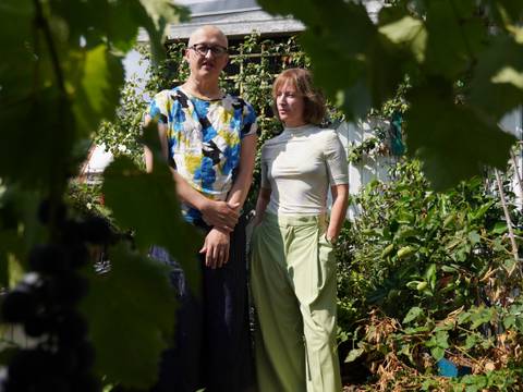  – Zwei Personen stehen in einem Garten, umgeben von Pflanzen. Eine trägt ein bunt gemustertes T-Shirt und eine Brille, die andere eine hellgrüne Hose und ein