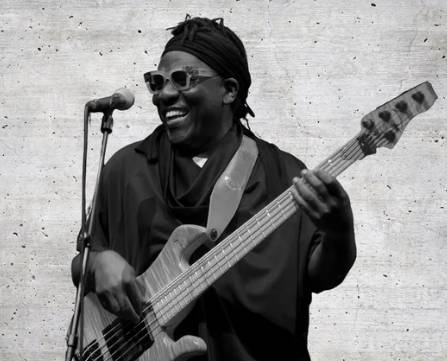 Richard Bona. Photo: Collins T. Photography – Person mit Sonnenbrille spielt E-Bass und lächelt. Mikrofon links, Hintergrund aus Beton.
