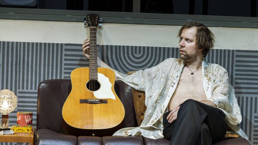 Stereophonic – Ein männlich gelesener Mensch auf einem Sofa hält eine Gitarre. Neben ihm steht eine Lampe und eine Dose. Hintergrund mit geometrischem Muster.