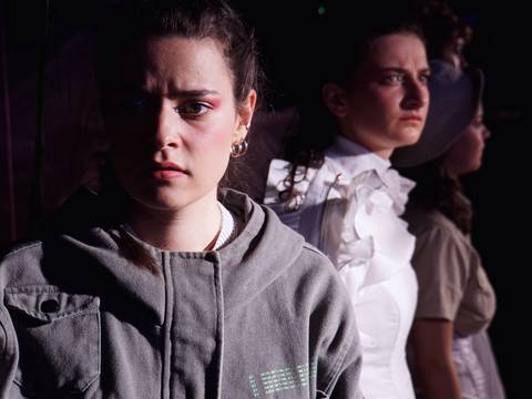  – Drei Personen stehen hintereinander. Die vorderste trägt eine graue Jacke, schaut nachdenklich. Die mittlere Person trägt ein weißes Oberteil mit Rüschen.