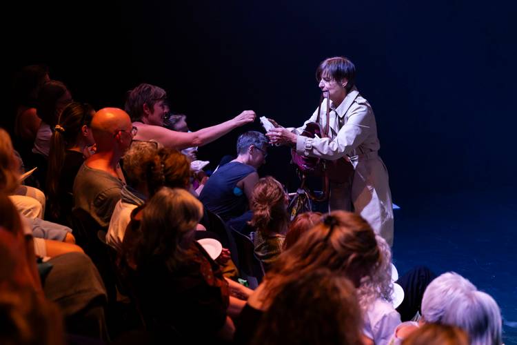 Person im Mantel reicht im dunklen Raum einem sitzenden Publikum Papiere oder Gegenstände. Links sind mehrere sitzende Personen zu sehen.
