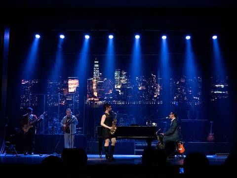  – Fünf Personen auf einer Bühne, eine Person spielt Saxophon, eine Klavier. Hintergrundbild einer beleuchteten Skyline. Bühnenscheinwerfer leuchten.