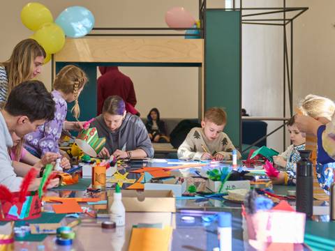  – Kinder und Erwachsene basteln am Tisch mit buntem Papier und Federn. Auf dem Tisch stehen Bastelmaterialien. Im Hintergrund hängen bunte Luftballons.