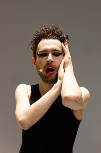 Future is Now – Person mit Make-up, rotem Lippenstift, schwarzem Lidschatten, Mikrofon am Gesicht, trägt ein ärmelloses schwarzes Oberteil, hält Gesicht mit beiden Händen.