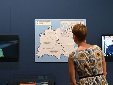  – Erwachsene Person von hinten betrachtet zwei Monitore und eine Karte mit Text: "Französischer Sektor", "Britischer Sektor", "Amerikanischer Sektor"