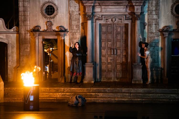 La forza del destino – Bühne mit zwei Personen; eine trägt einen Mantel, die andere ein glitzerndes Kleid; zwischen ihnen eine Tür; brennendes Fass links im Vordergrund.