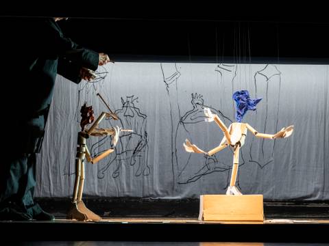 Die Geschichte vom Soldaten, Salzburger Marionettentheater – Zwei Holzkasper stehen auf einer Bühne, ein Mensch bedient die Fäden, Hintergrund Leinwand mit Zeichnungen.