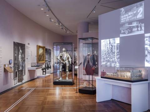  – Raumansicht „Objekte. Geschichte. Geschichten. Blick in die Sammlung“. Im Vordergrund auf der rechten Seite ist eine Vitrine mit einem Modell des Zeughauses vor einer Projektionsfläche zu sehen. Auf dieser sind mehrere Schwarz-Weiß-Fotos zu erkennen. Im Hintergrund sind verschiedene Ausstellungsobjekte in Vitrinen zu sehen, darunter eine Ritterrüstung und eine Militäruniform. An der Wand auf der linken Seite hängen Gemälde und Fotos.