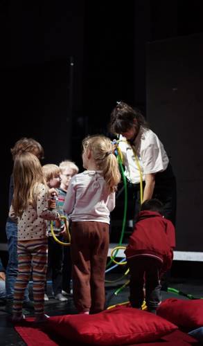 Knirpskonzerte – Kinder stehen um eine Person mit Schlaeuchen. Kissen liegen auf dem Boden. Die Kinder tragen gemusterte Kleidung, die Person ein weisses Hemd.