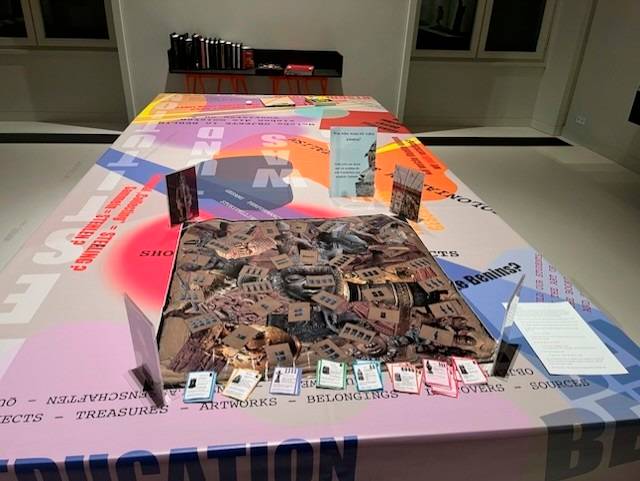 Decolonial Gaming: The Talking Benin Bronzes bei der Langen Nacht der Museen 2026 – Bunter Tisch mit Aufdrucken wie EDUCATION, ARTWORKS, und einem zentralen Bild. Verschiedene Broschüren und Infotafeln umrahmen das Bild.