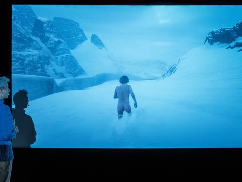  – Person in blauem Pullover sieht auf Bildschirm mit unbemalter Person, die durch schneebedeckte Landschaft geht, links große Berge.