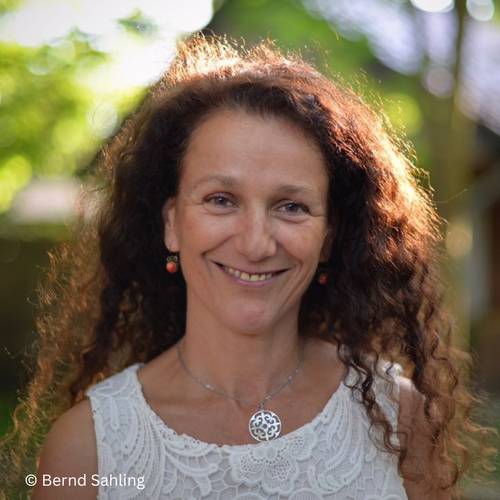 Anja Tuckermann – Eine lächelnde Person mit lockigem Haar trägt ein weißes Spitzenoberteil und eine silberne Halskette. Text: © Bernd Sahling.