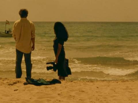  – Ein Mann und eine Frau stehen am Strand und schauen auf das Meer, die Frau, rechts im Bild, hält eine Filmkamera in der Hand.