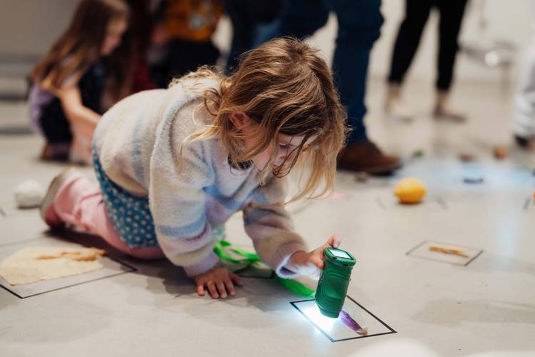 Foto: Ein Kind sitzt mit Taschenlampe zwischen kreativem Material im Ausstellungsraum auf dem Boden.