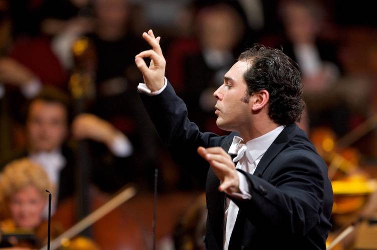 Tugan Sokhiev – Dirigent mit dunklem Haar und Anzug dirigiert ein Orchester mit erhobenen Händen in einem Konzertraum. Verschwommene Musiker*innen im Hintergrund.
