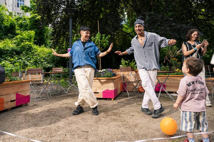 Jhow und Gissauro führen im Freien eine Hip-Hop-Tanzszene mit offenen Armen auf, während ein Publikum aus Kindern und Erwachsenen aufmerksam zuschaut und mitklatscht.