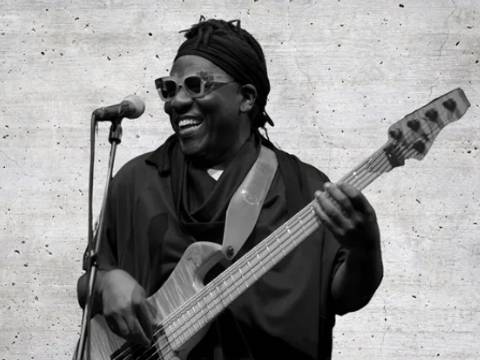 Richard Bona. Photo: Collins T. Photography – Person mit Sonnenbrille spielt E-Bass und lächelt. Mikrofon links, Hintergrund aus Beton.