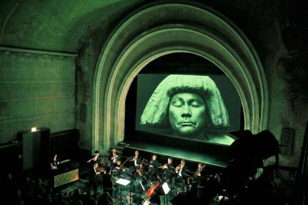 GOLEM – Orchester spielt in einem Theater mit hohem Bogen; große Leinwand zeigt Skulptur mit Augen geschlossen, Stirnband und langen Haaren.