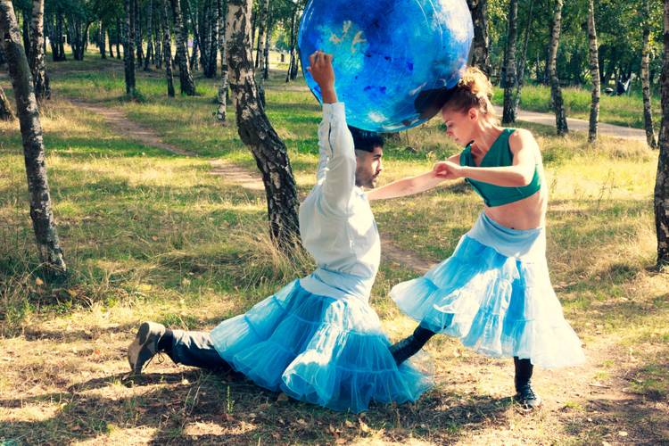 Zwei Personen im Wald. Eine hält einen großen blauen Globus über ihrem Kopf. Beide tragen blaue Röcke. Die Sonne scheint durch die Bäume.