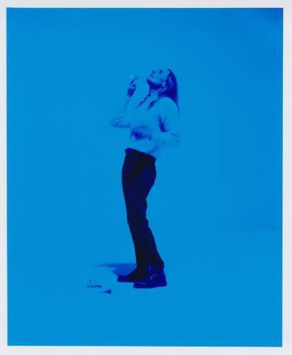 Foto: Estelle Hanania – Person in blauem Licht, trägt Pullover und Hose, blickt nach oben, hält Gegenstand in der Hand; Schatten und Objekte auf dem Boden.