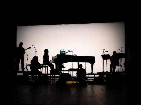  – Schatten von sechs Personen mit Musikinstrumenten auf einer Bühne vor hellem Hintergrund, darunter ein Klavier und fünf Mikrofone auf Ständern.