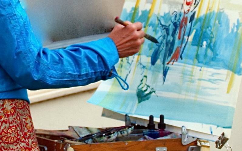 Person mit Pinsel vor einer bereits – vorwiegend blau – bemalten Leinwand