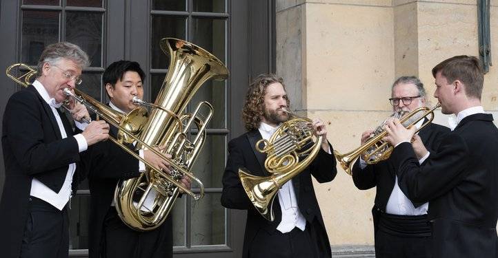 Konzerthaus Brass Berlin – Fünf Personen in schwarzen Anzügen und Fliegen spielen Blechblasinstrumente vor einem Gebäude mit Steinfassade und großen Fenstern.