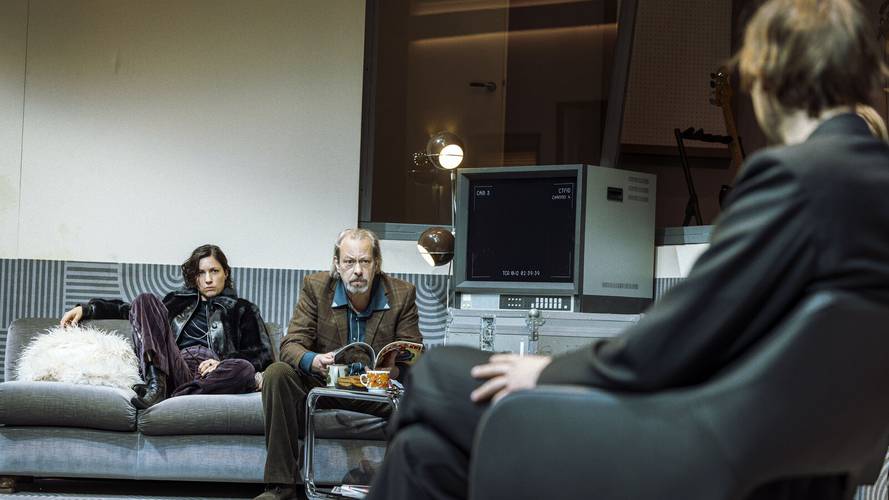 Stereophonic – Zwei Personen sitzen auf einem Sofa, eine dritte Person sitzt im Vordergrund. Ein Bildschirm zeigt "CAM 8 CH0001". Ein Tisch mit Tassen steht vor ihnen.