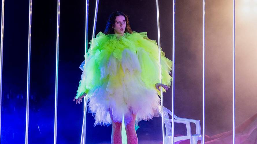 Person in fluoreszierendem Tüllkleid steht zwischen leuchtenden LED-Streifen; Hintergrund dunkel, seitlich ein weißer Stuhl sichtbar.