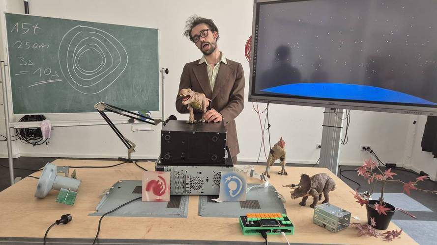 Person im Anzug mit Spielzeugdinosaurier in der Hand steht vor Tisch mit Computerteilen und Dinosauriern. Tafel mit Kreisen und Text: 15t, 250m, 3s, = 10s.