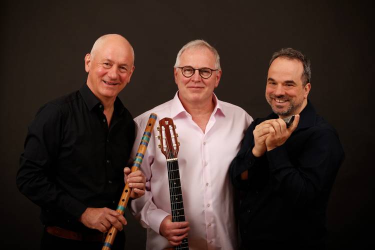 Drei männlich gelesene Personen mit Instrumenten: Eine hält eine Flöte, eine eine Gitarre und eine eine Mundharmonika. Alle lächeln in die Kamera.