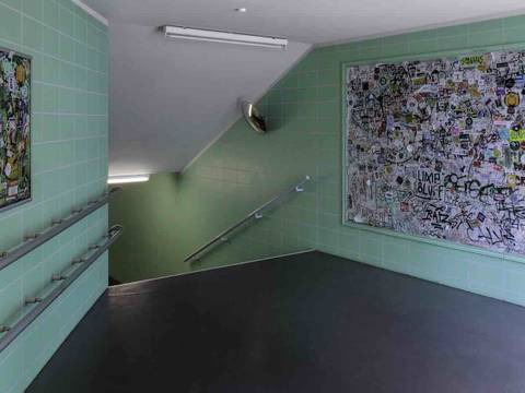 Robert Kusmirowski, Transition (Übergang), 2009 fortwährend, Staatliche Museen zu Berlin, Nationalgalerie – Grüngraue gekachelte Wand mit drei gerahmten Flächen, voll mit bunten Stickern und Graffiti. Ein Durchgang führt in eine Treppe abwärts.