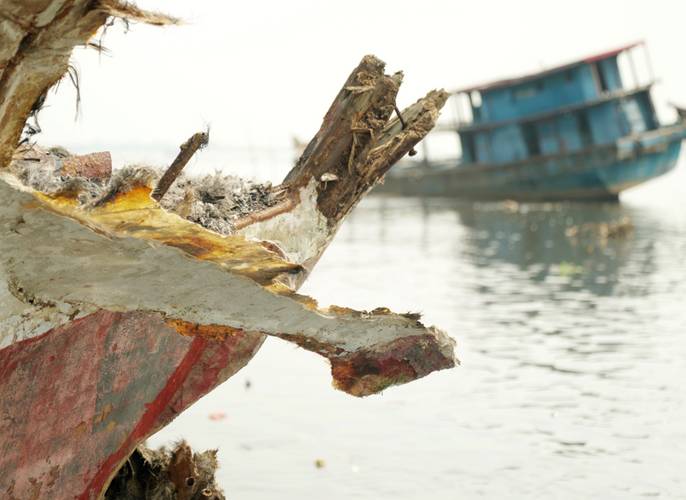 Ocean Bound 3 – Zerbrochenes Holzboot im Vordergrund, unscharfes blaues Boot im Hintergrund auf ruhigem Wasser.