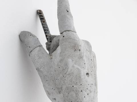 Auf Gute Nachbarschaft. – Graue Betonskulptur einer Hand an der Wand, die ein Metallstab durchbohrt. Einige Finger sind gestreckt.