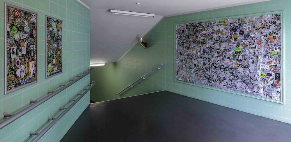 Robert Kusmirowski, Transition (Übergang), 2009 fortwährend, Staatliche Museen zu Berlin, Nationalgalerie – Grüngraue gekachelte Wand mit drei gerahmten Flächen, voll mit bunten Stickern und Graffiti. Ein Durchgang führt in eine Treppe abwärts.