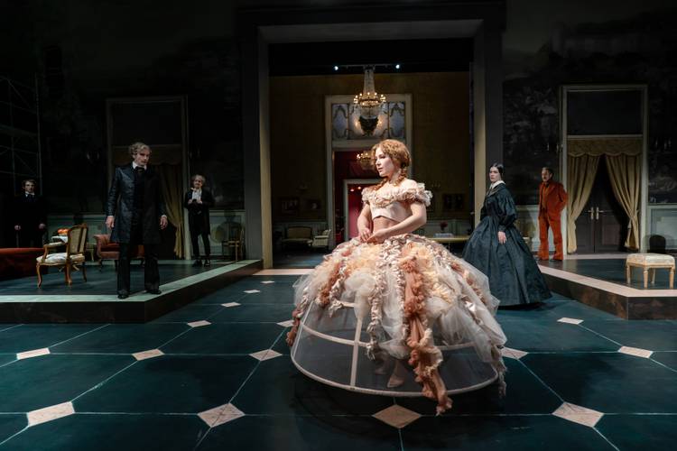 Il Gattopardo – Eine junge Frau in einem Reifrock steht in einem Salon, im Hintergrund sind weitere Personen in historischen Kostümen