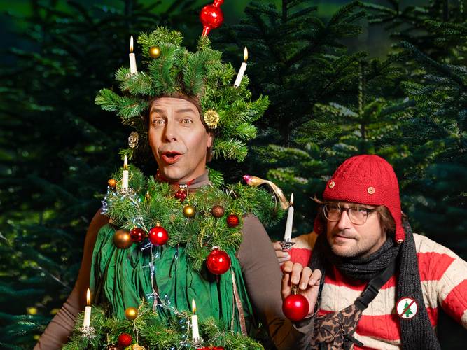 Märchen im Glaspalast von und mit dem Hexenberg Ensemble: Das Mädchen mit den Schwefelhölzern | Der Tannenbaum – Ein als Weihnachtsbaum verkleidete Person mit Baumschmuck und Kerzen neben einer rot-weiß gestreift gekleideten Person mit roter Mütze vor Tannenbäumen.